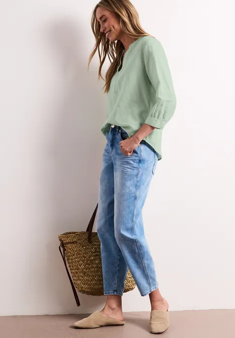 Dame trägt grüne Bluse, lockere blaue Jeans, beige Slipper und hält einen Korb in der Hand.